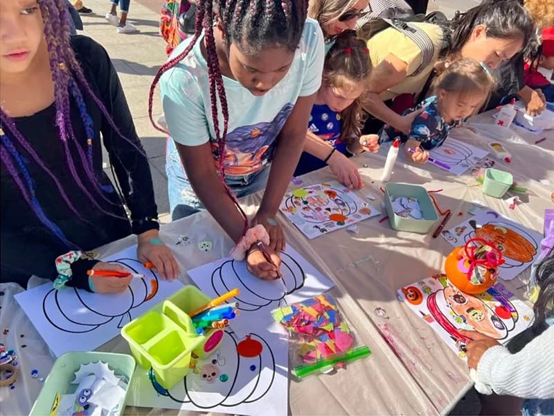 Kids and parents decorating paper pumpkins at a craft table during an outdoor autumn event