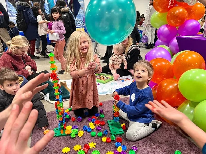 Young children playing with building toys on the floor at an indoor family event, a balloon arch behind them